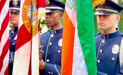 Three police officers stand holding the USA flag far left, the Florida flag center, and the Coral Gables flag on the right