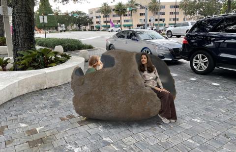 Digitally added large brown stone sits on the plaza and two women, photoshopped, are whispering to one another