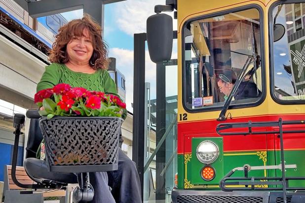On sunny day with clouds, a woman with a green shirt sits on her wheelchair smiling in front of the trolley