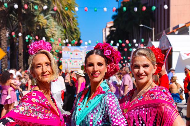 Three women dressed in colorful flamenco shawls and flower headpieces smile for the camera at Feria de Sevilla.