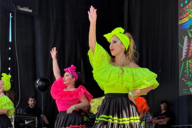 Two women in traditional Colombian green and pink dresses dance with one arm raised. Spectators sit in the front with hats in the crowd