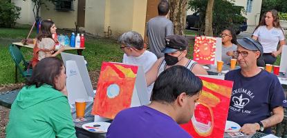Adults sit at a picnic table and paint small canvases 