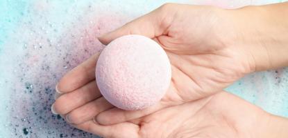 Two hands hold a spherical bath bomb over light blue, soapy water