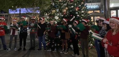 At night under the light of a Christmas tree, teenage carolers in red and green attire with Santa hats sing angelic songs.