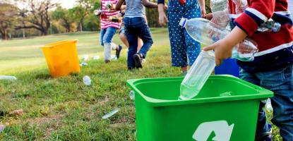 Children pick up plastic water bottles from the grass. One child places them in a green recycling bin.