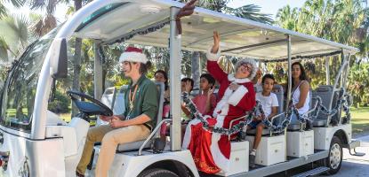 Employee with elf ears drives a large golf cart with Santa and passengers around a field of palm trees on a warm and sunny day