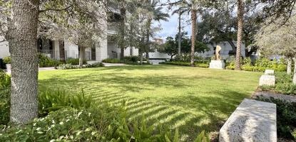 A tree shades a stone seating and flowers and sun hits an open space lawn, a sculpture in the distance