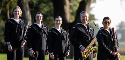 Five musicians in dark blue Navy uniform hold wind instruments and stand on a field of grass. In a shallow depth background is a large tree and open field.