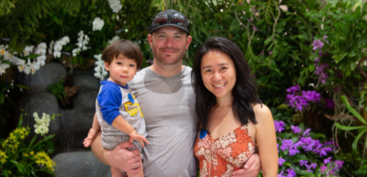 A toddler, father, and mother smile for photo in front of a floral back drop