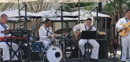 Four men in white, Navy uniform play instruments in the shade on a sunny day.