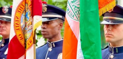 Three police officers stand holding the USA flag far left, the Florida flag center, and the Coral Gables flag on the right