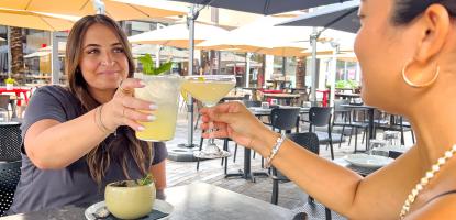 Two women sitting down clink their cocktails together on sunny day