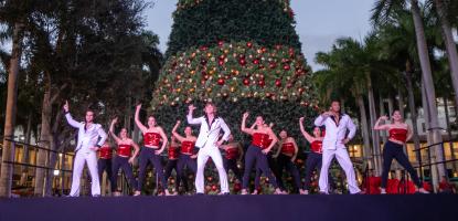 3 singers in white at the front of the stage and dancers in red behind them. Behind them an enormous Christmas tree