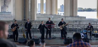 Outside stage by the water Navy band in blue uniform play on stage, in the foreground are audience members