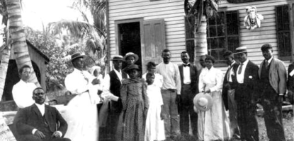 Historic black-and-white photo of early settlers in Coral Gables, showing a group of men, women, and children dressed in formal attire standing outside a wooden house with palm trees.
