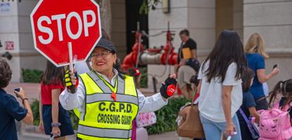 Crossing Guard in neon green vest holds a stop sign, in the background children cross the street carrying and pulling backpacks