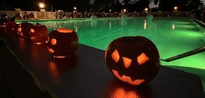 Carved pumpkins gloat in the dark night by a lit pool