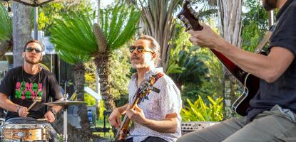 Guitarists play under a tent with sunlight bleeding through on green plants