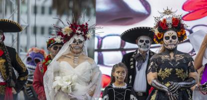 People in elaborate Día de los Muertos costumes and face paint stand on stage, including a bride in a white gown, a woman in a black and gold dress with skeleton body paint, mariachi-style performers, and a child in a skeleton dress.