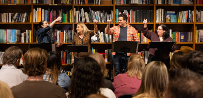 Actors performing in front of a bookcase, in the foreground audience members sit and watch