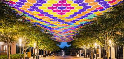 Colorful crocheted canopy in purple, yellow, orange, pink, and green stretches above  tree-lined pedestrian (Giralda Plaza) walkway illuminated at dusk.