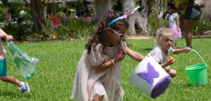 Little girl in bunny ears and sunglasses grabs plastic egg on a green lawn with other kids running