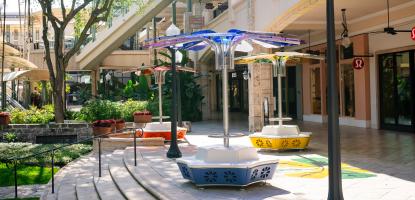 Flower art installations with seating are displayed on shady and sunny shops plaza with a stores and a tree in the background