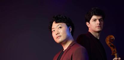 Two musicians stand in a dark background with their stringed instruments
