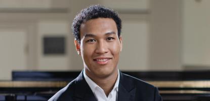 Alt text  Pianist Clayton Stephenson sits in front of a grand piano, wearing a black suit and white shirt.