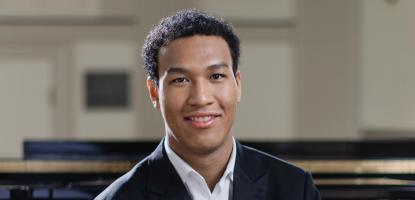 Alt text  Pianist Clayton Stephenson sits in front of a grand piano, wearing a black suit and white shirt.