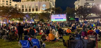 Photo of people sitting in lawn chairs and towels on grass watching a soccer game on a large monitor