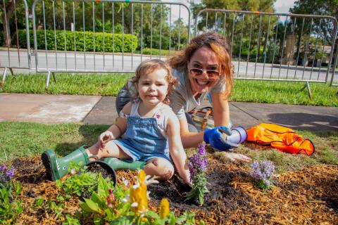 A warm sunny day, a toddler and mother sit on the grass and dirt with recently planted plants with purple flowers.