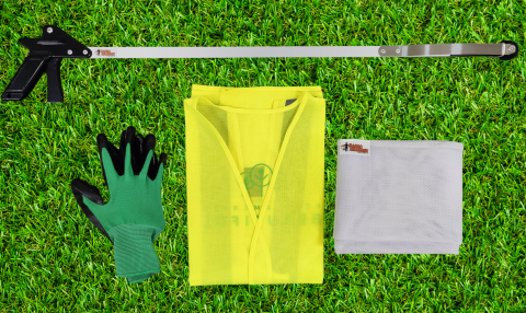 On a background of green grass, a green glove, yellow vest in the middle, and garbage net lie. A garbo grabber lies above the other items. 