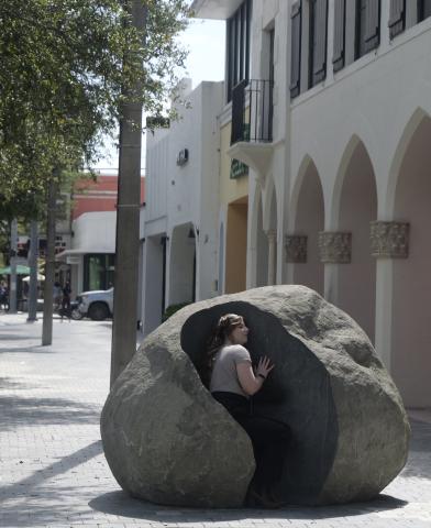 A woman sits inside a large rock sculpture with her ear pressed against it on a hot sunny day