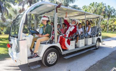 Employee with elf ears drives a large golf cart with Santa and passengers around a field of palm trees on a warm and sunny day