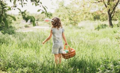 A child stands in a grassy field facing away from the camera. They are wearing bunny ears and holding a basket of colorful eggs
