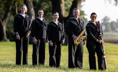 Five musicians in dark blue Navy uniform hold wind instruments and stand on a field of grass. In a shallow depth background is a large tree and open field.