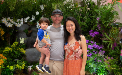 A toddler, father, and mother smile for photo in front of a floral back drop