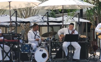 Four men in white, Navy uniform play instruments in the shade on a sunny day.