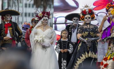 People in elaborate Día de los Muertos costumes and face paint stand on stage, including a bride in a white gown, a woman in a black and gold dress with skeleton body paint, mariachi-style performers, and a child in a skeleton dress.