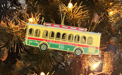 Yellow, red, and green Coral Gables trolley as an ornament hanging on a tree with white lights