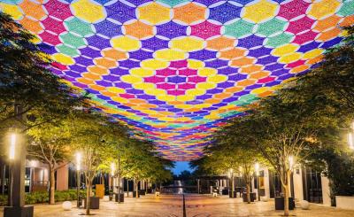 Colorful crocheted canopy in purple, yellow, orange, pink, and green stretches above  tree-lined pedestrian (Giralda Plaza) walkway illuminated at dusk.
