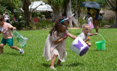 Little girl in bunny ears and sunglasses grabs plastic egg on a green lawn with other kids running