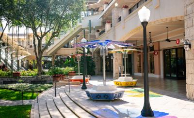 Flower art installations with seating are displayed on shady and sunny shops plaza with a stores and a tree in the background