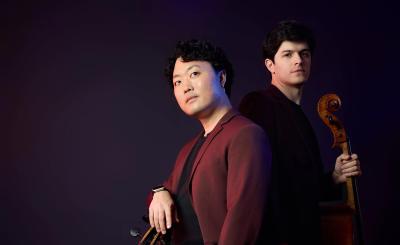 Two musicians stand in a dark background with their stringed instruments