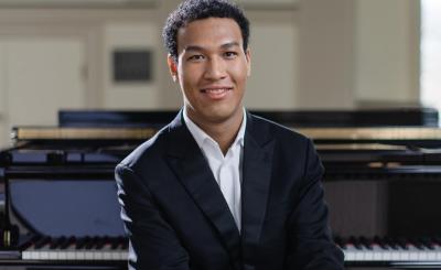 Alt text  Pianist Clayton Stephenson sits in front of a grand piano, wearing a black suit and white shirt.