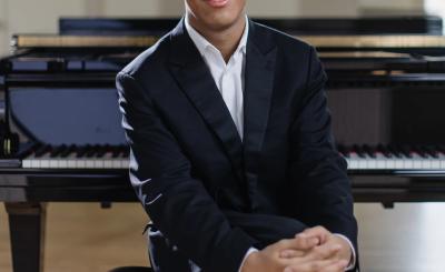 Alt text  Pianist Clayton Stephenson sits in front of a grand piano, wearing a black suit and white shirt.