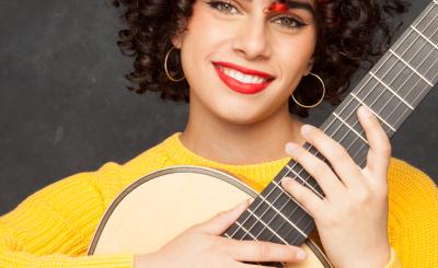 Guitarist Leonela Alejandro smiles while holding a classical guitar and wearing a yellow sweater.