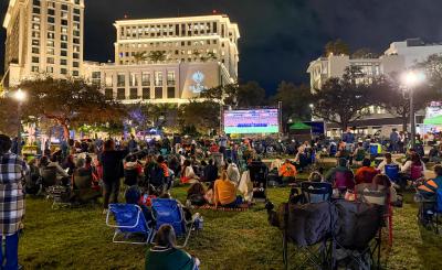 Photo of people sitting in lawn chairs and towels on grass watching a soccer game on a large monitor