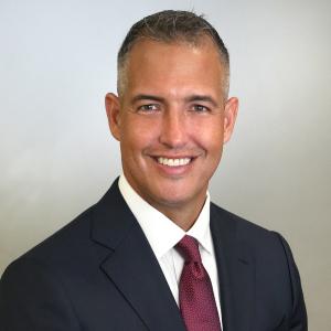Billy Urquia in dark suit smiling against white/gray background